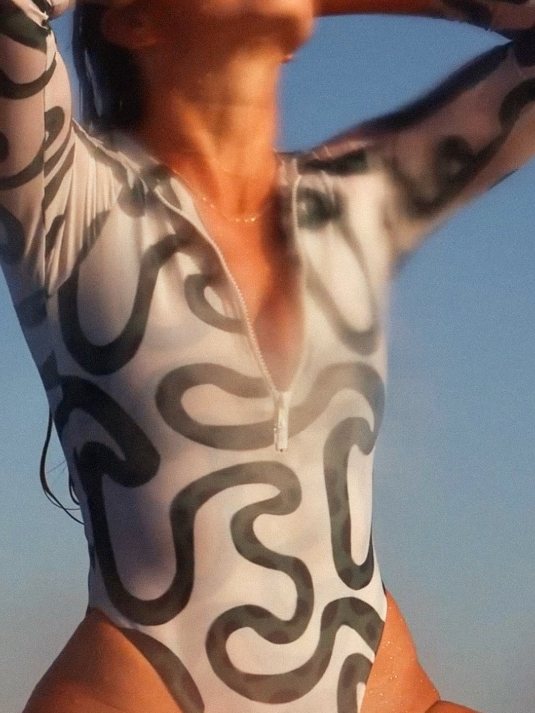 Person wearing a surf bodysuit with green and white abstract patterns against a clear blue sky.