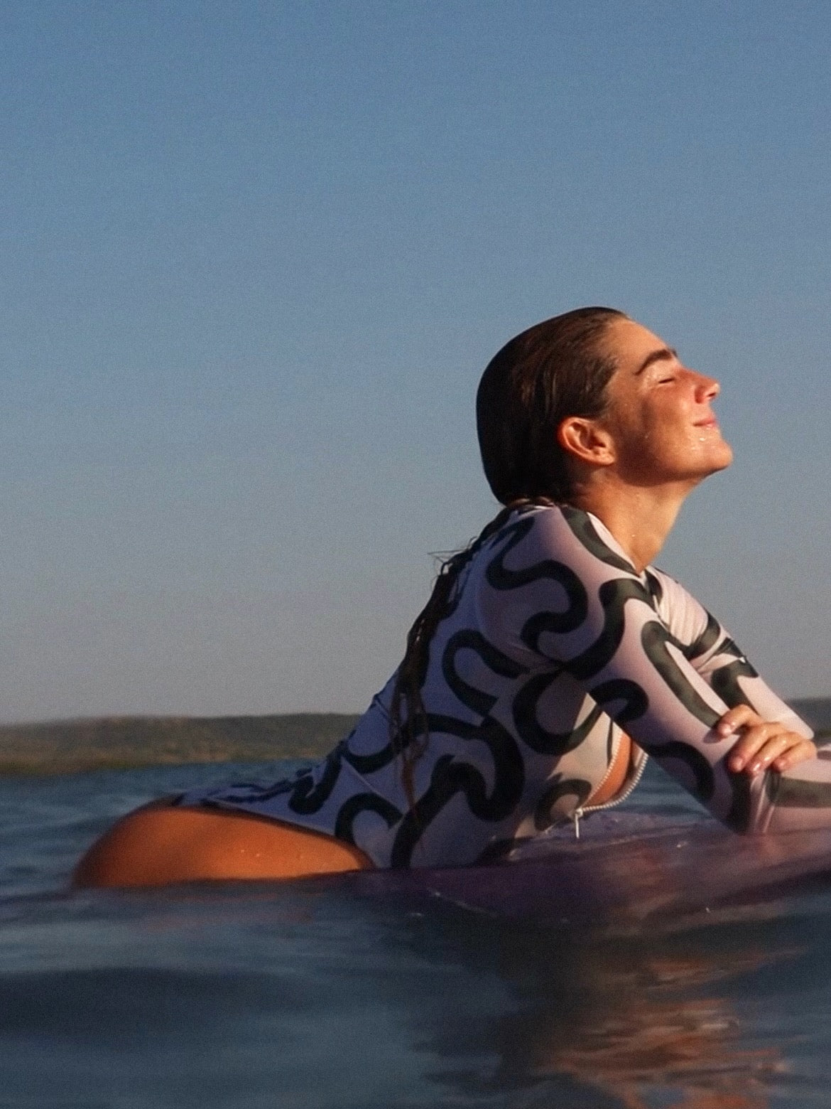 Person in a green and white abstract patterned swimsuit sitting on a surfboard in the ocean with a clear blue sky.